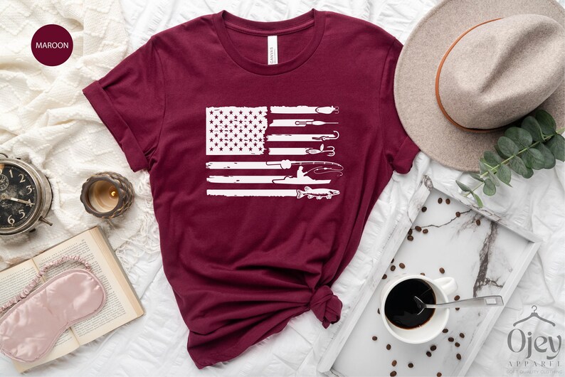 K&ouml;nnte beinhalten: Ein maroonfarbenes T-Shirt mit einem wei&szlig;en, abgenutzten Design einer amerikanischen Flagge mit Angelruten, K&ouml;dern und einem Fisch. Die Flagge ist mittig auf dem Shirt platziert.