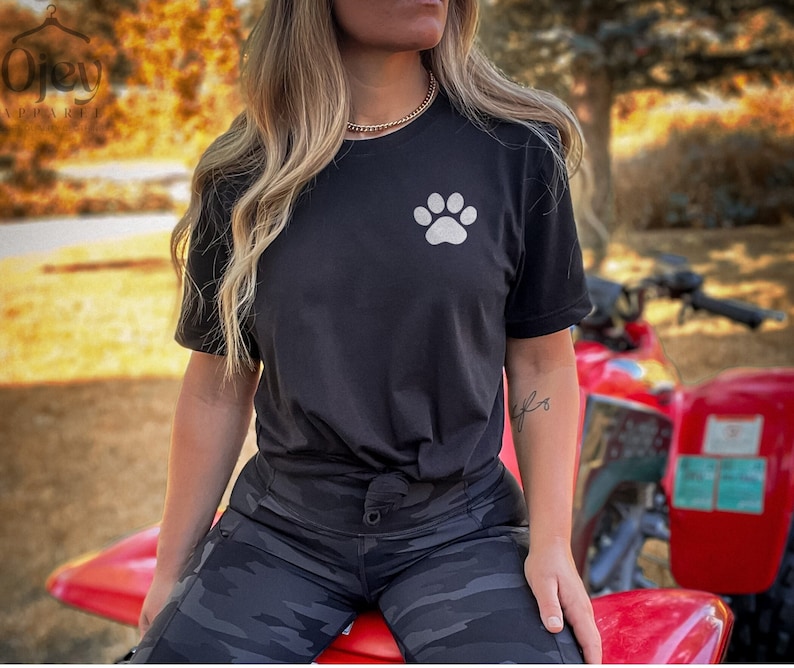 May include: A black t-shirt with a white paw print design. The shirt is being worn by a person with long blonde hair. The person is sitting on a red ATV.