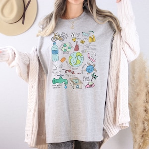 May include: A gray t-shirt with a colorful graphic design featuring various environmental messages and illustrations, including a globe, a turtle, a tree, a faucet, and the words "Recycle, Reduce, Reuse, Save the Planet, Don't Litter, Save Water, Save Life, Plant a Tree".