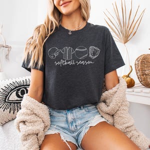 May include: A woman wearing a dark grey t-shirt with a white graphic of softball equipment and the text "softball season". She is wearing blue denim shorts and a white fuzzy cardigan.
