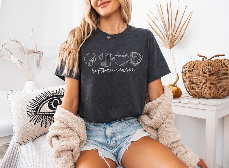 May include: A woman wearing a dark grey t-shirt with a white graphic of softball equipment and the text "softball season". She is wearing blue denim shorts and a white fuzzy cardigan.