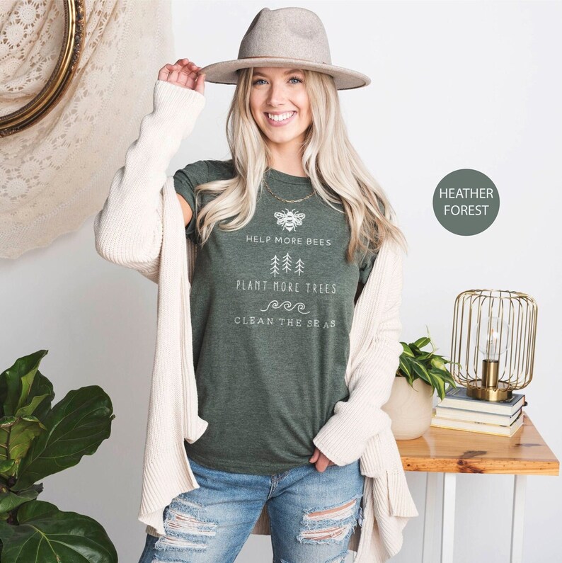K&ouml;nnte beinhalten: Ein gr&uuml;nes T-Shirt mit dem Text "Help More Bees, Plant More Trees, Clean The Seas" und einer Bienengrafik. Das Shirt wird von einer Person mit blonden Haaren und einem braunen Hut getragen.