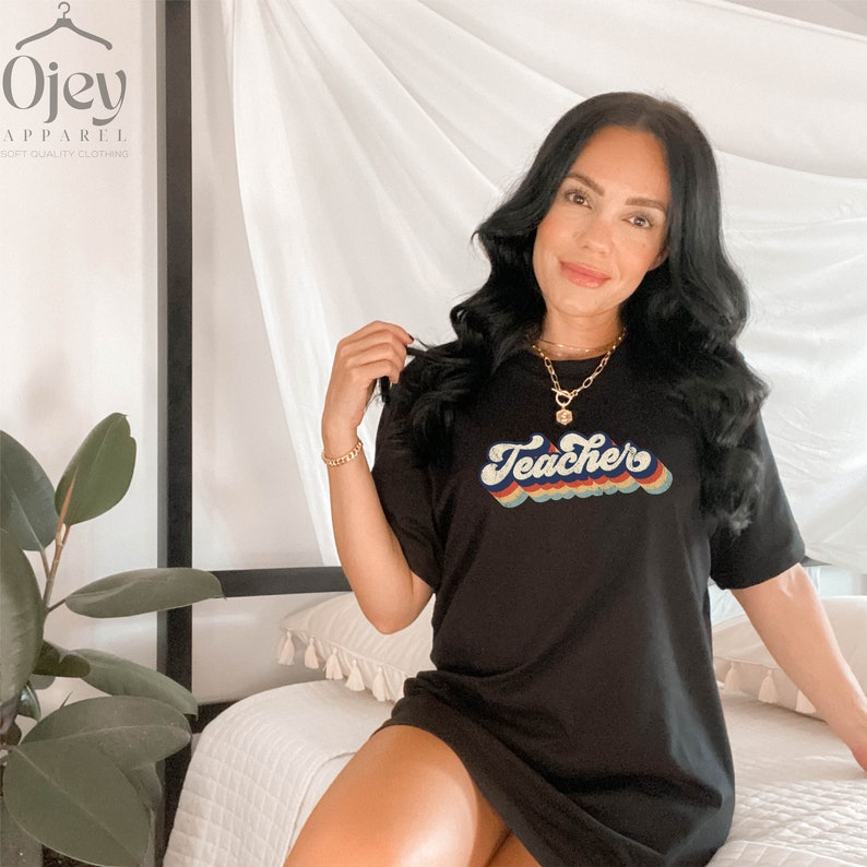 Puede incluir: Un vestido de camiseta negro con un gr&aacute;fico "Teacher" de colores arco&iacute;ris en una fuente retro.