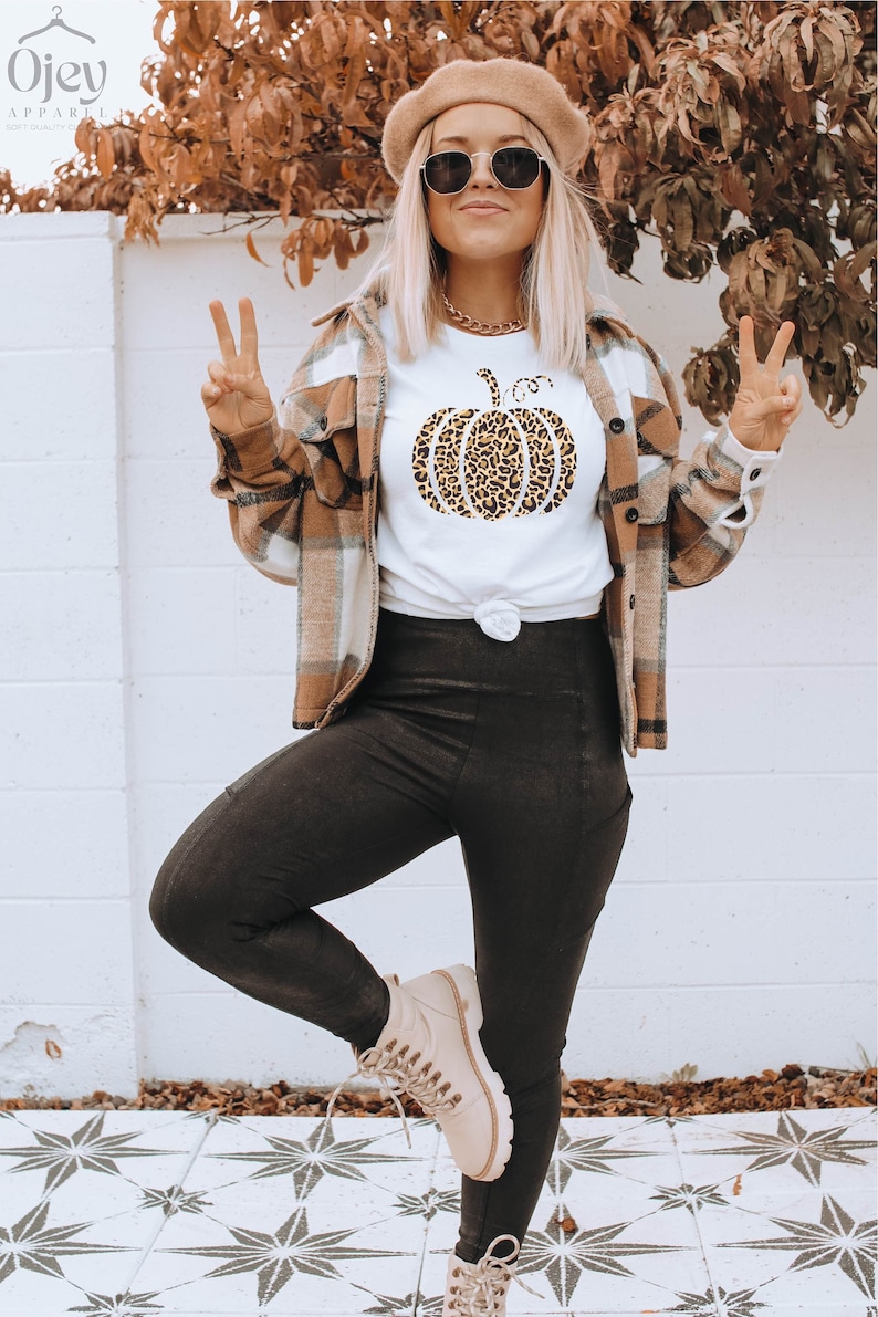 May include: A white t-shirt with a leopard print pumpkin graphic. The model is wearing black leggings and brown boots. She is also wearing a brown and white plaid shirt and a brown beret.