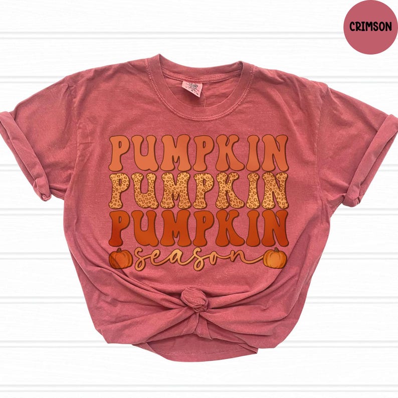 May include: Crimson-colored t-shirt with the words "PUMPKIN PUMPKIN PUMPKIN season" in orange and leopard print lettering. Two small pumpkins are on the bottom of the shirt. The shirt is knotted at the bottom.
