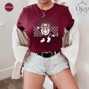May include: A maroon t-shirt with a white checkered background and a smiling baseball character wearing a hat. The text "GAMEDAY" is above the character.