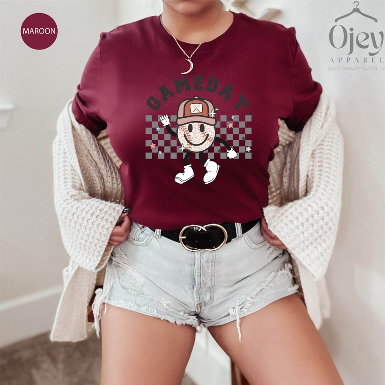 May include: A maroon t-shirt with a white checkered background and a smiling baseball character wearing a hat. The text "GAMEDAY" is above the character.