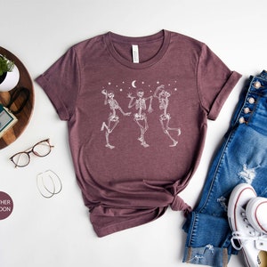 May include: A heather maroon t-shirt featuring a white graphic of three skeletons dancing beneath a crescent moon and stars. The shirt is laid flat on a white surface with a pair of glasses, a wooden tray with a camera, and a pair of white trainers and blue jeans.
