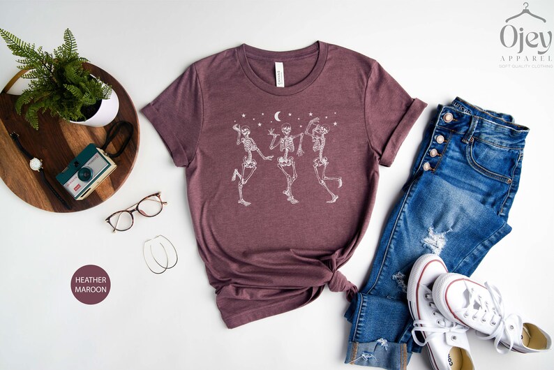 May include: A heather maroon t-shirt featuring a white graphic of three skeletons dancing beneath a crescent moon and stars. The shirt is laid flat on a white surface with a pair of glasses, a wooden tray with a camera, and a pair of white trainers and blue jeans.