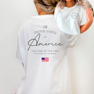 Peut inclure: T-shirt blanc avec un imprim&eacute; graphique des mots "The United States of America" et le texte "The Land of the Free, The Home of the Brave". Le graphique comprend &eacute;galement un petit drapeau am&eacute;ricain. L'avant du t-shirt a un petit graphique d'un cercle avec des &eacute;toiles et le nombre "1776" &agrave; l'int&eacute;rieur.
