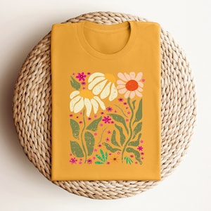 May include: A mustard yellow t-shirt with a floral print featuring white, pink, and green flowers. The flowers are stylized and have a retro feel.