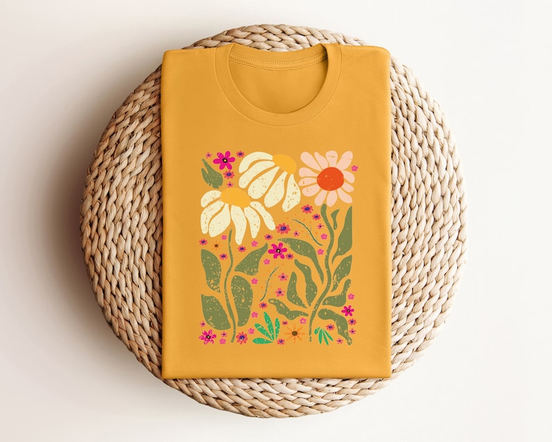 May include: A mustard yellow t-shirt with a floral print featuring white, pink, and green flowers. The flowers are stylized and have a retro feel.