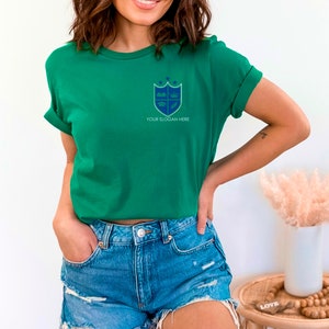 May include: A vibrant green t-shirt featuring a blue and white crest design. The crest includes a book, crown, and leaves, with the text "YOUR SLOGAN HERE" beneath. The model is wearing blue denim shorts.