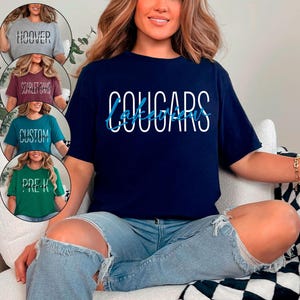 以下が含まれることがあります： 白で「COUGARS」、青で「Lakeview」と書かれたネイビーブルーのTシャツ。画像には、「HOOVER」、「SCARLET OAKS」、「CUSTOM」、「PRE-K」という言葉が書かれた他の4つのTシャツも表示されています。