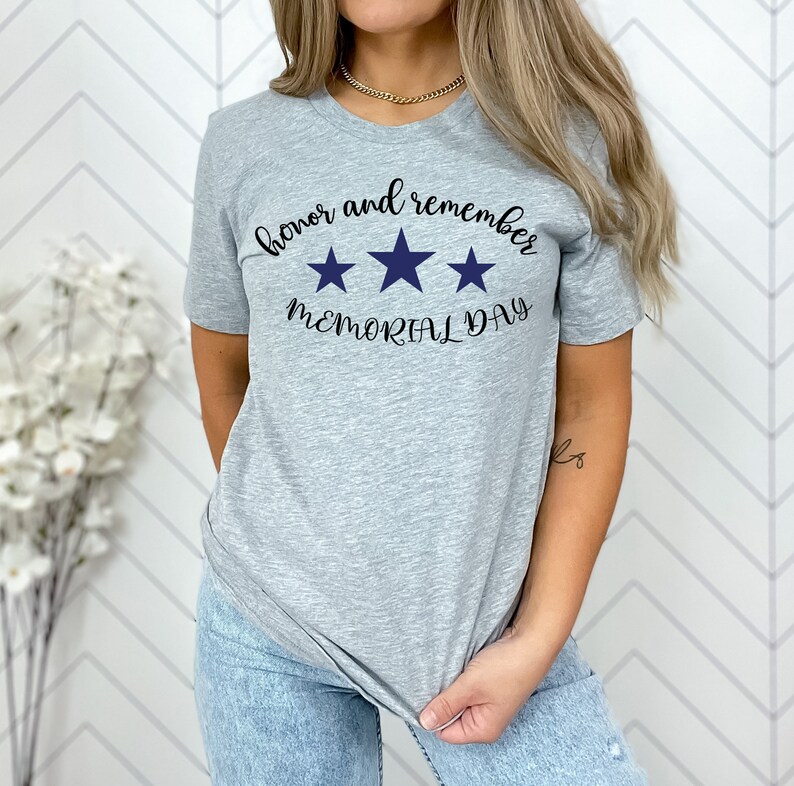 以下が含まれることがあります： 黒い文字で「honor and remember MEMORIAL DAY」と書かれたグレーのTシャツ。テキストの上に青い星が3つある。