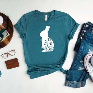 May include: A teal heather deep teal t-shirt with a white silhouette of a rabbit with floral details. The t-shirt is laid flat on a white surface with a pair of jeans and white sneakers.