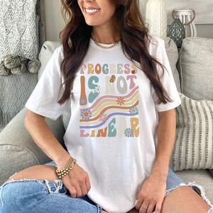 May include: White t-shirt with a graphic design that says "Progress is not linear" in a retro font with colorful flowers and wavy lines.