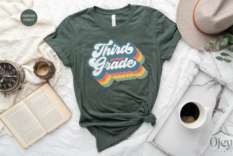 May include: A green t-shirt with the text "Third Grade" in a retro rainbow design.