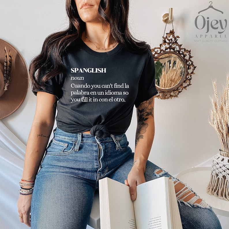 Puede incluir: Camiseta negra con texto blanco que dice "SPANGLISH noun Cuando you can't find la palabra en un idioma so you fill it in con el otro."