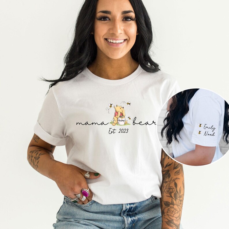May include: White t-shirt featuring a Winnie the Pooh illustration and the text "mama bear Est. 2023". The shirt also has a small bee illustration on the sleeve with the text "Emily Noah".