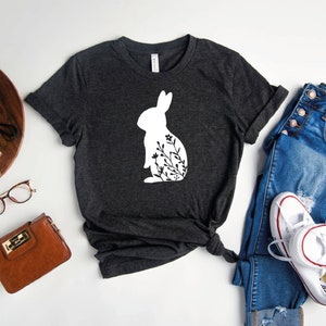 May include: A dark heather gray t-shirt with a white silhouette of a rabbit with floral details. The shirt is laid flat on a white surface with a pair of jeans, a purse, and a pair of glasses.