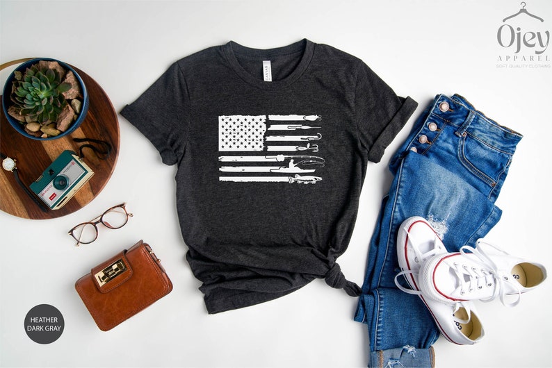 K&ouml;nnte beinhalten: Ein dunkelgraues T-Shirt mit einem wei&szlig;en, abgenutzten Design einer amerikanischen Flagge mit Werkzeugen. Die Flagge besteht aus Werkzeugen wie Schraubenschl&uuml;sseln, H&auml;mmern und Schraubendrehern.
