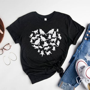 May include: A black t-shirt with a white silhouette of a heart made up of cats. The t-shirt is laid out on a white surface with a pair of jeans, a pair of white sneakers, a camera, a watch, and a pair of glasses.