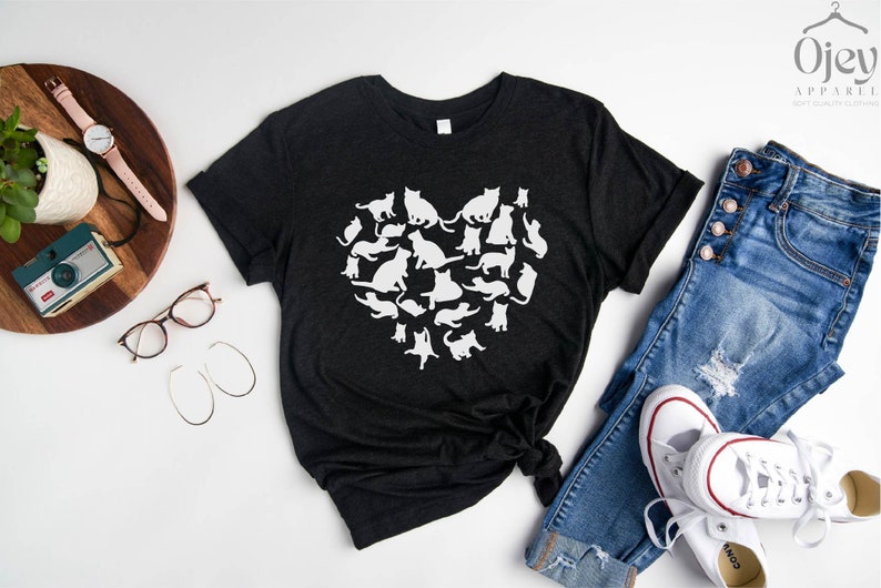 May include: A black t-shirt with a white silhouette of a heart made up of cats. The t-shirt is laid out on a white surface with a pair of jeans, a pair of white sneakers, a camera, a watch, and a pair of glasses.
