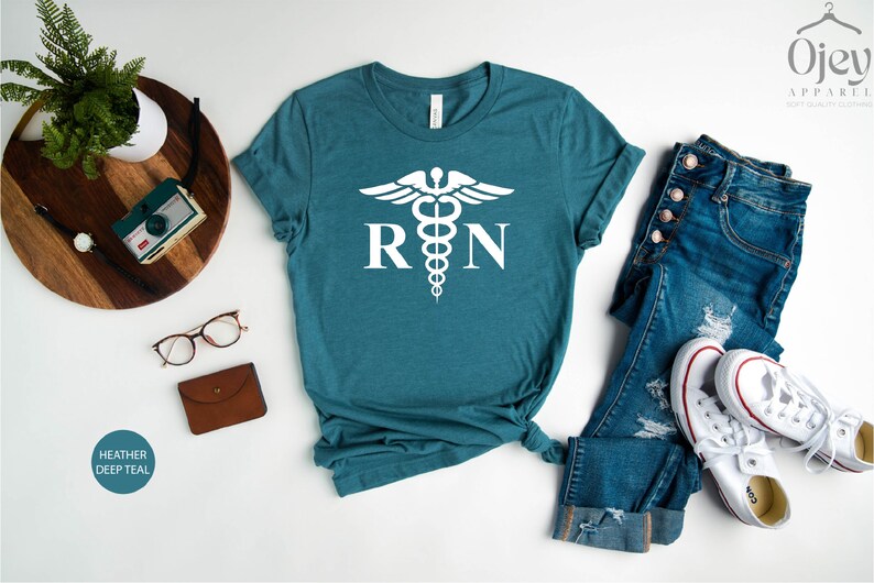May include: A heather deep teal t-shirt with a white medical caduceus symbol and the letters "RN" printed on the front. The shirt is laid flat on a white surface with a pair of jeans, a wallet, a pair of glasses, and a camera.