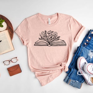 K&ouml;nnte beinhalten: Ein T-Shirt in der Farbe Heidepfirsich mit einer schwarzen Grafik eines offenen Buches, aus dem Blumen wachsen. Das Shirt liegt auf einer wei&szlig;en Oberfl&auml;che mit einem Buch, einer Brille, einer Geldb&ouml;rse und einer Jeans und wei&szlig;en Turnschuhen.