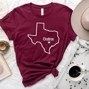 Op de afbeelding: Maroon T-shirt met een witte omtrek van de staat Texas en het woord "Dallas" binnen de omtrek. Een klein wit hartje bevindt zich in het midden van de staat.