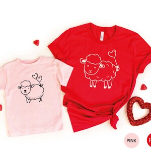 Peut inclure: Deux t-shirts avec un motif de mouton mignon. Le t-shirt rose a un mouton noir avec deux c&oelig;urs qui sortent de sa queue. Le t-shirt rouge a un mouton blanc avec un ballon en forme de c&oelig;ur attach&eacute; &agrave; sa queue. Les t-shirts sont sur un fond blanc avec des c&oelig;urs rouges.