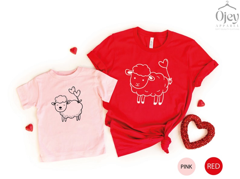 Peut inclure: Deux t-shirts avec un motif de mouton mignon. Le t-shirt rose a un mouton noir avec deux c&oelig;urs qui sortent de sa queue. Le t-shirt rouge a un mouton blanc avec un ballon en forme de c&oelig;ur attach&eacute; &agrave; sa queue. Les t-shirts sont sur un fond blanc avec des c&oelig;urs rouges.