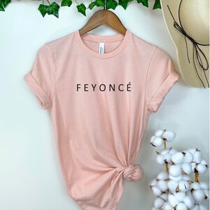 Pu&ograve; includere: Una maglietta rosa chiaro con la scritta "FEYONC&Eacute;" stampata in nero sul davanti.