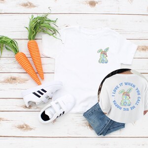 Puede incluir: Camiseta blanca con un conejito arco&iacute;ris con un sombrero en la parte delantera y el texto "Me encanta cuando me llamas big hoppa" en la parte posterior.
