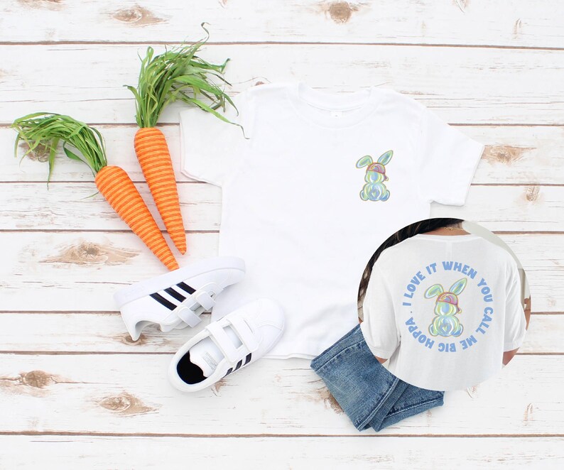 Puede incluir: Camiseta blanca con un conejito arco&iacute;ris con un sombrero en la parte delantera y el texto "Me encanta cuando me llamas big hoppa" en la parte posterior.