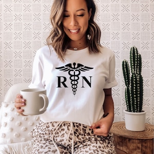 May include: White t-shirt with a black medical caduceus symbol and the letters "RN" printed on the front. The person is wearing leopard print pants.