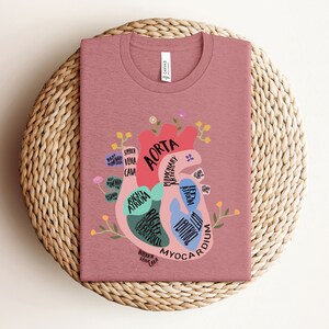 May include: A pink t-shirt with a graphic of a human heart with anatomical labels. The heart is surrounded by flowers and leaves. The text on the heart includes "Aorta", "Right Atrium", "Right Ventricle", "Left Atrium", "Left Ventricle", "Pulmonary Artery", "Superior Vena Cava", "Inferior Vena Cava", and "Myocardium".