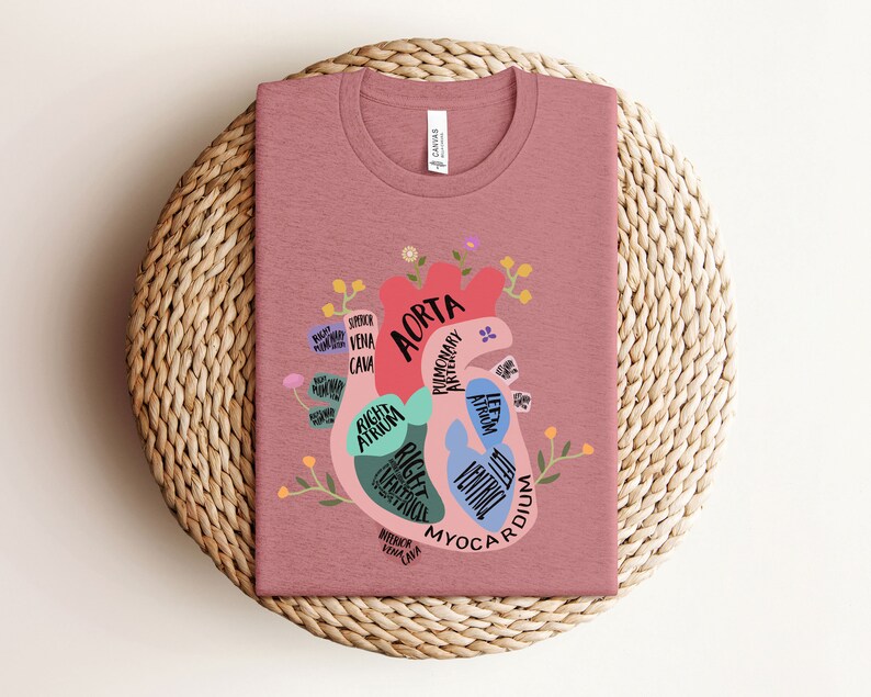 May include: A pink t-shirt with a graphic of a human heart with anatomical labels. The heart is surrounded by flowers and leaves. The text on the heart includes "Aorta", "Right Atrium", "Right Ventricle", "Left Atrium", "Left Ventricle", "Pulmonary Artery", "Superior Vena Cava", "Inferior Vena Cava", and "Myocardium".
