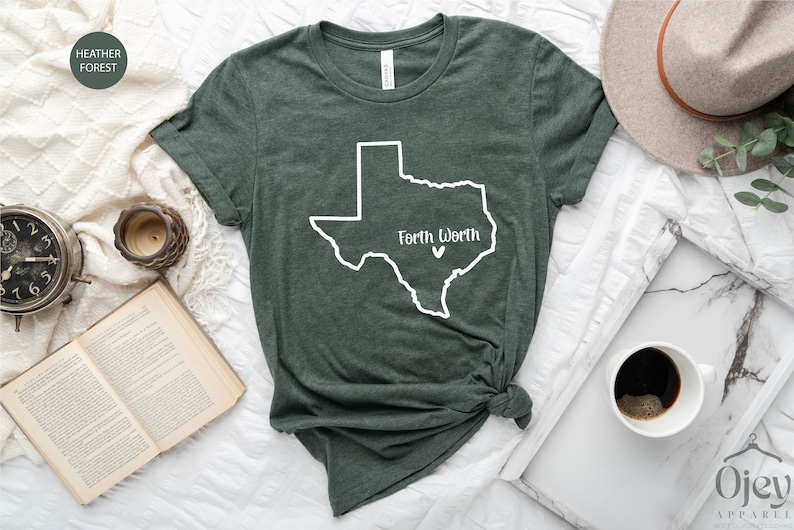Op de afbeelding: Een donkergroen t-shirt met een witte omtrek van de staat Texas en de tekst "Fort Worth" met een klein wit hartje binnen de omtrek van de staat.