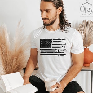 K&ouml;nnte beinhalten: Ein wei&szlig;es T-Shirt mit einem schwarzen und wei&szlig;en, abgenutzten Design einer amerikanischen Flagge mit Fischereimotiven. Die Flagge zeigt eine Silhouette einer Person beim Angeln, einen Fisch und Angelausr&uuml;stung.