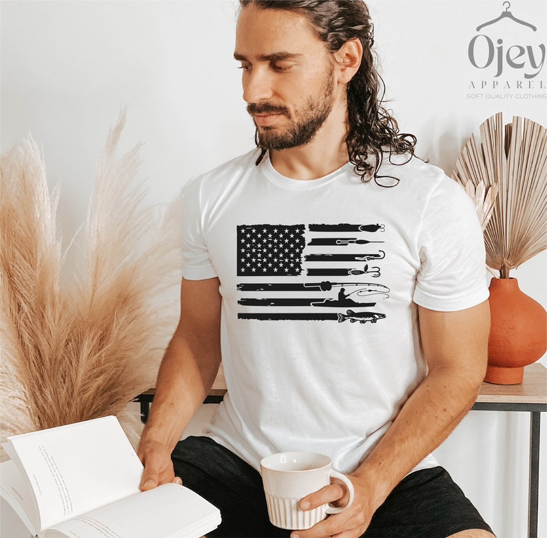 K&ouml;nnte beinhalten: Ein wei&szlig;es T-Shirt mit einem schwarzen und wei&szlig;en, abgenutzten Design einer amerikanischen Flagge mit Fischereimotiven. Die Flagge zeigt eine Silhouette einer Person beim Angeln, einen Fisch und Angelausr&uuml;stung.