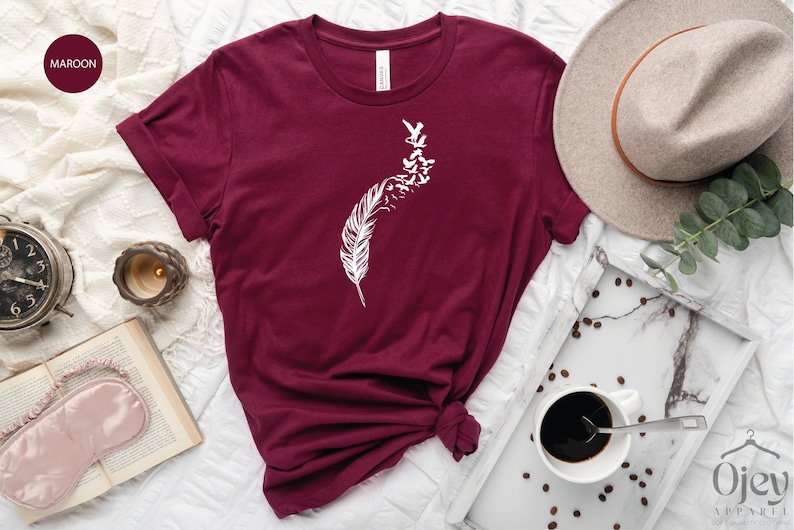 Op de afbeelding: Een maron t-shirt met een witte afbeelding van een veer met vogels die er vanaf vliegen.