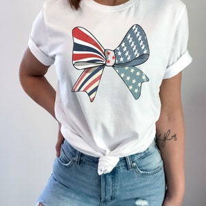 May include: A white t-shirt with a red, white, and blue bow design. The bow is made up of two overlapping pieces of fabric, one with red and white stripes and the other with blue and white stars. The bow is centered on the shirt and is slightly off-center.