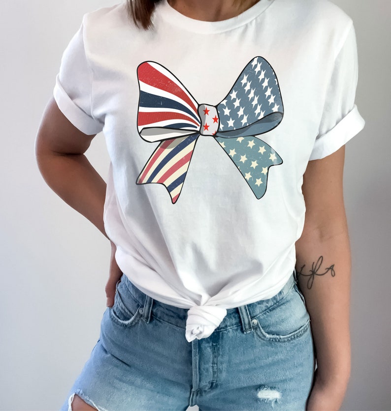 May include: A white t-shirt with a red, white, and blue bow design. The bow is made up of two overlapping pieces of fabric, one with red and white stripes and the other with blue and white stars. The bow is centered on the shirt and is slightly off-center.