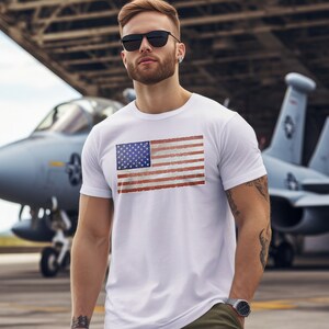 Pode incluir: Uma camiseta branca com um design de bandeira americana vintage. A bandeira est&aacute; desgastada e desbotada, o que lhe confere um aspeto desgastado. O modelo est&aacute; a usar a camiseta e est&aacute; em p&eacute; em frente a uma aeronave militar.
