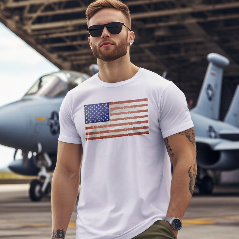Pode incluir: Uma camiseta branca com um design de bandeira americana vintage. A bandeira est&aacute; desgastada e desbotada, o que lhe confere um aspeto desgastado. O modelo est&aacute; a usar a camiseta e est&aacute; em p&eacute; em frente a uma aeronave militar.