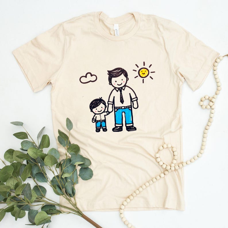 Peut inclure: Un t-shirt beige avec un dessin d'un p&egrave;re et de son fils se tenant la main. Le dessin est en noir, bleu et jaune. Le soleil brille et il y a un nuage dans le ciel.
