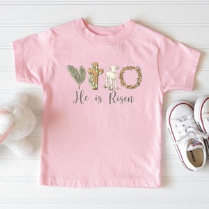 May include: Pink toddler t-shirt with a white lamb, cross, and palm branch graphic. The text "He is Risen" is printed below the graphic.