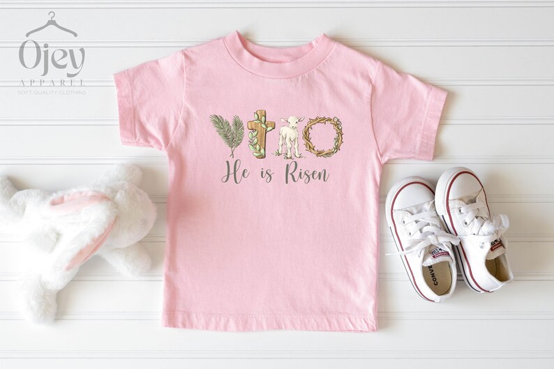 May include: Pink toddler t-shirt with a white lamb, cross, and palm branch graphic. The text "He is Risen" is printed below the graphic.